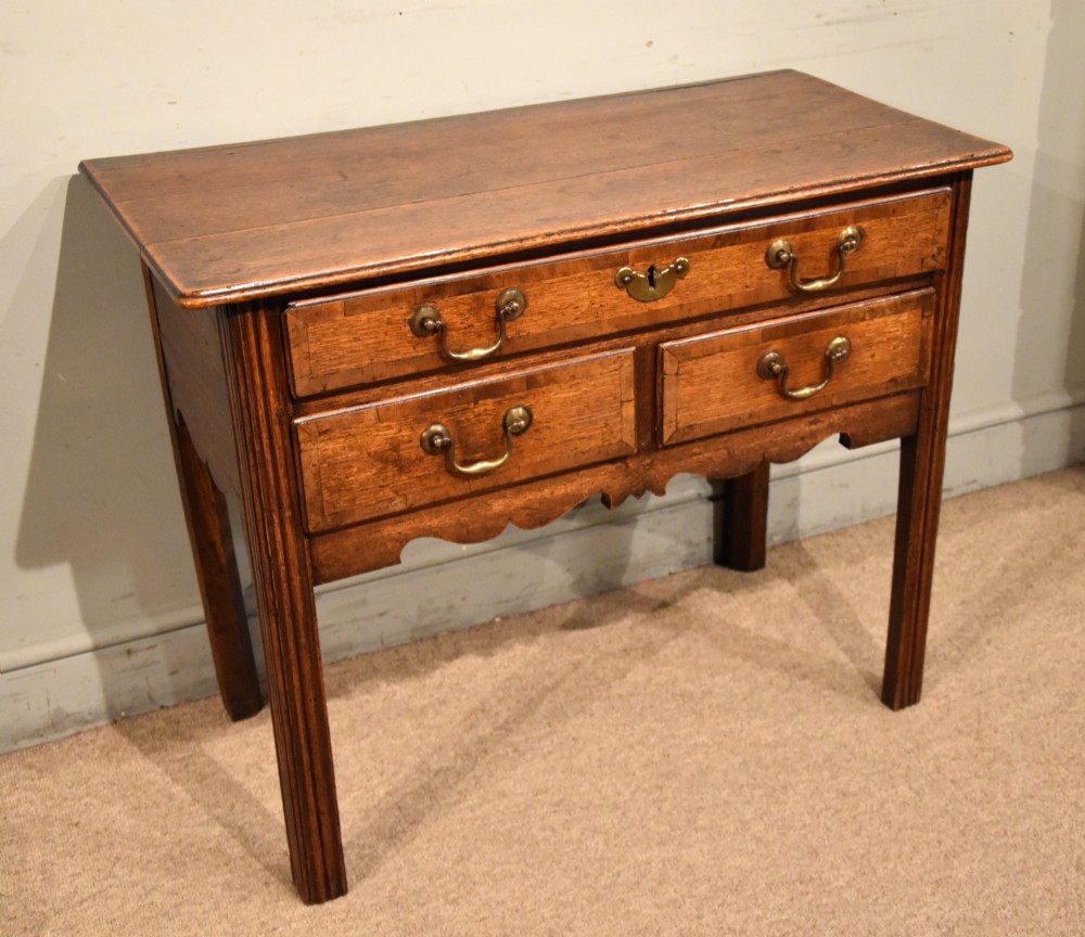 A George III oak lowboy side table featuring one wide upper drawer above two smaller drawers, fitted with brass swan-neck handles, a scalloped apron, and reeded square legs. View II