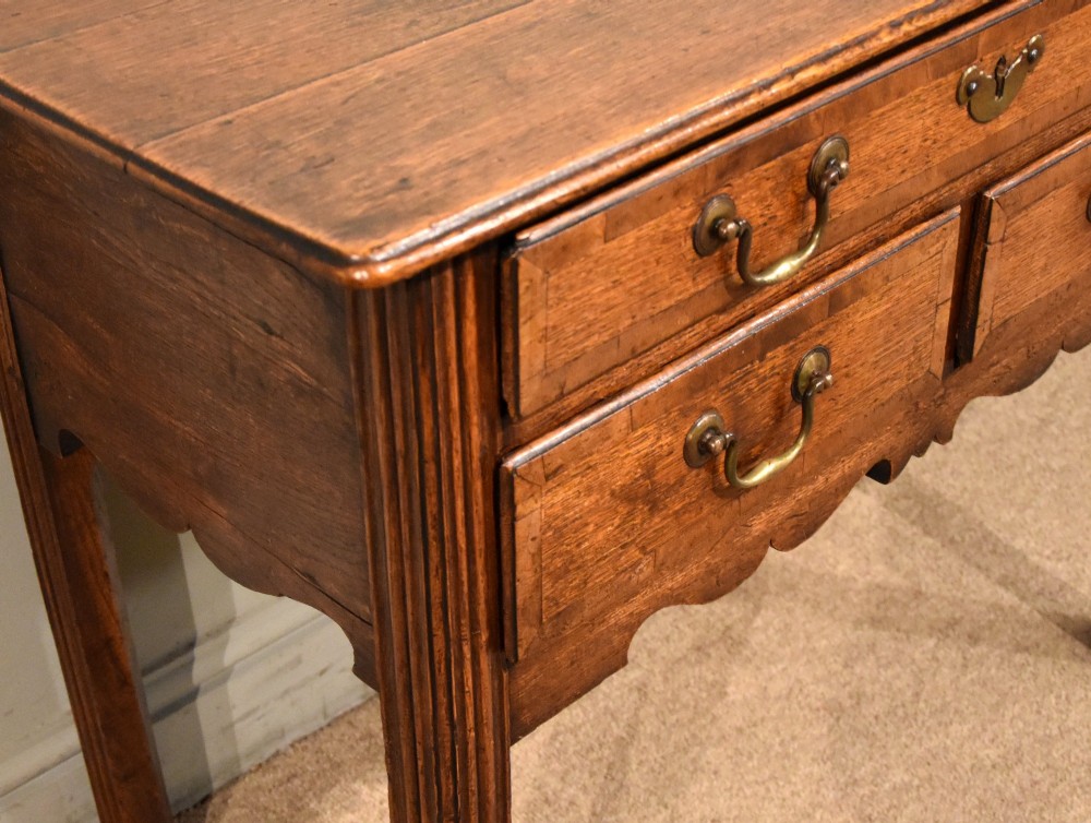 A George III oak lowboy side table featuring one wide upper drawer above two smaller drawers, fitted with brass swan-neck handles, a scalloped apron, and reeded square legs. View V