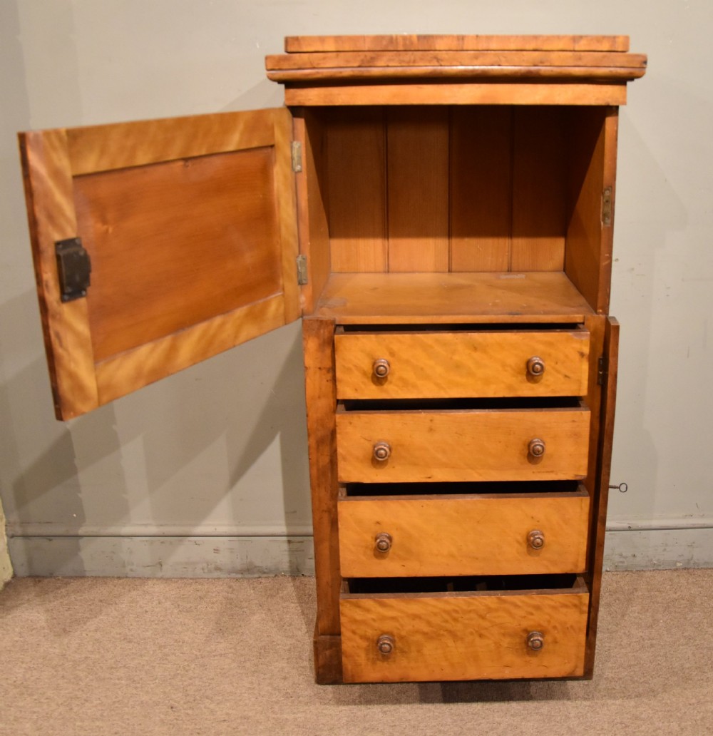 A tall Victorian satin birch Wellington chest featuring multiple drawers, a top secretaire compartment, turned wooden knobs, and a hinged side locking bar on a plinth base. View IV