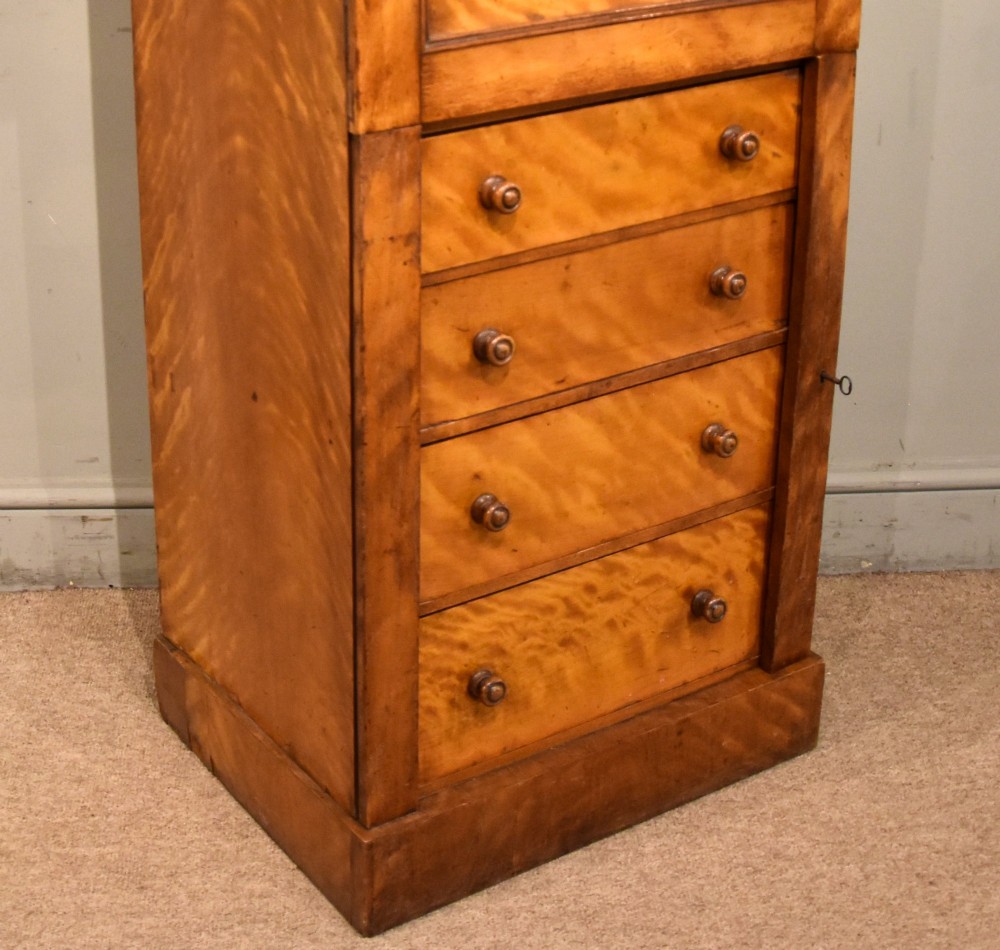 A tall Victorian satin birch Wellington chest featuring multiple drawers, a top secretaire compartment, turned wooden knobs, and a hinged side locking bar on a plinth base. View V