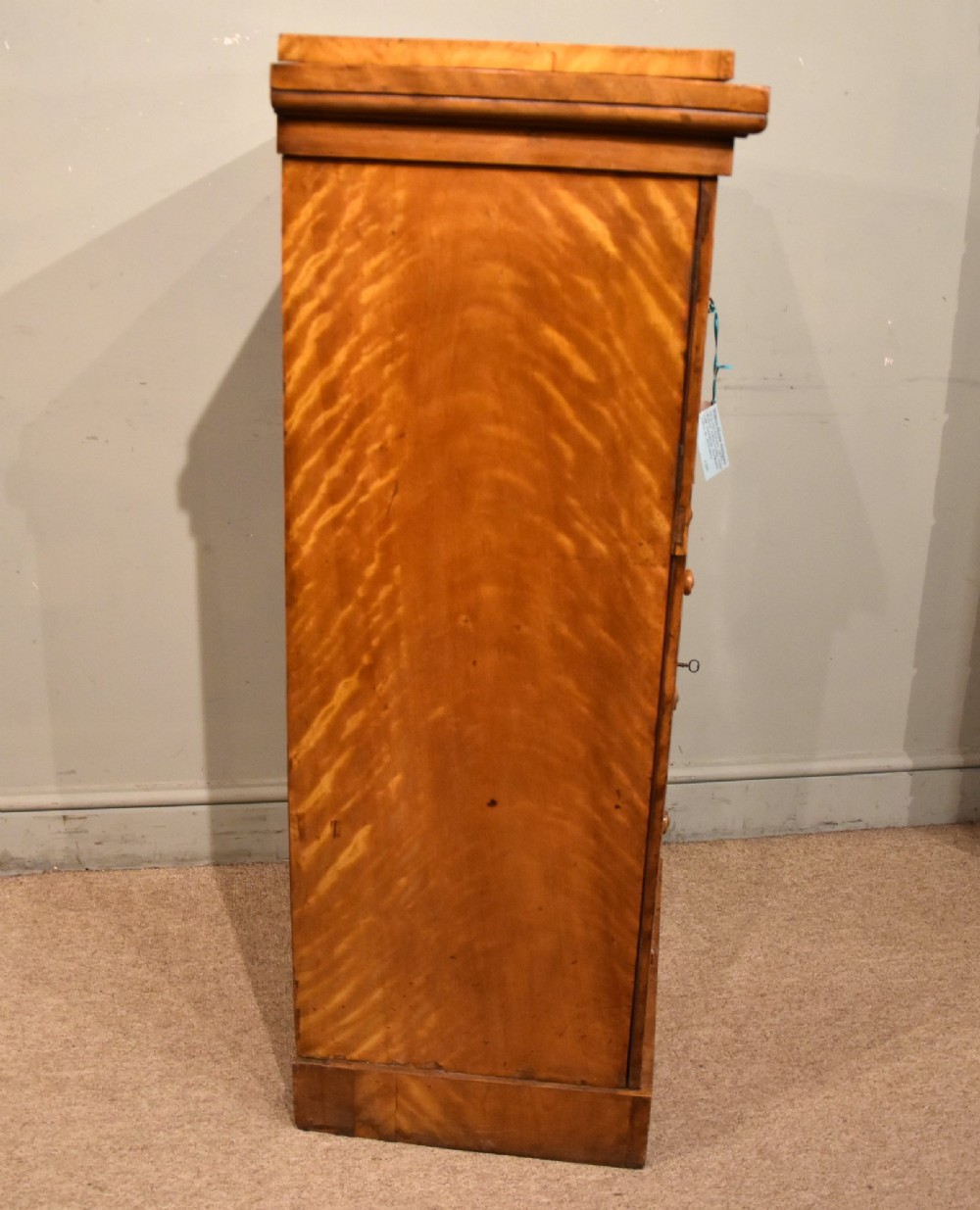 A tall Victorian satin birch Wellington chest featuring multiple drawers, a top secretaire compartment, turned wooden knobs, and a hinged side locking bar on a plinth base. View VI