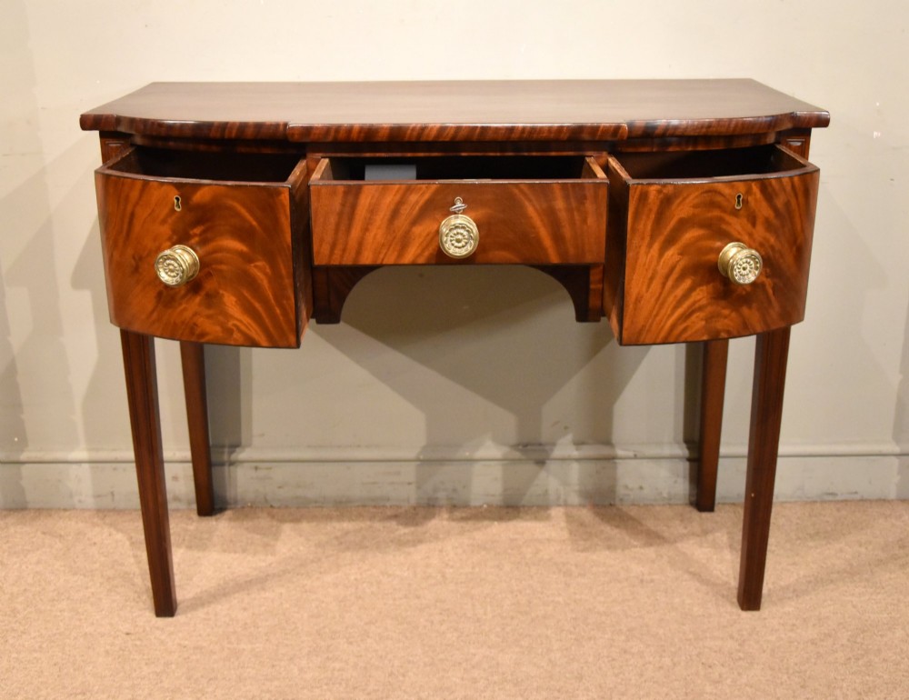 A Regency period flame mahogany bowfront sideboard featuring a central drawer flanked by two curved doors, set on tapered square legs with round brass handles. View II