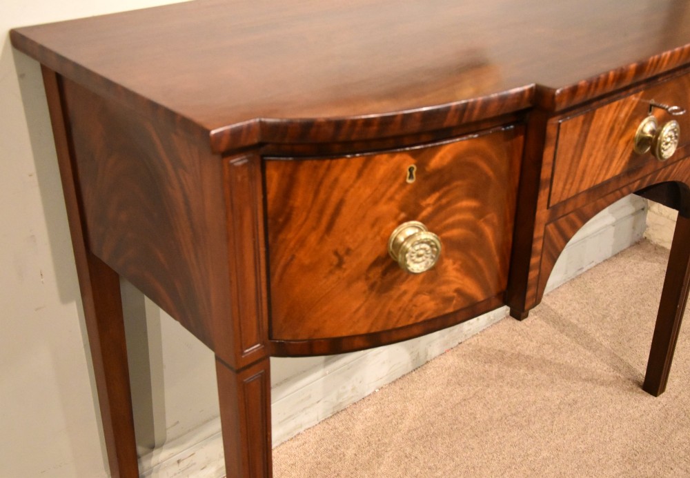 A Regency period flame mahogany bowfront sideboard featuring a central drawer flanked by two curved doors, set on tapered square legs with round brass handles. View V