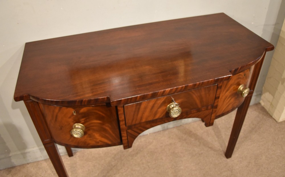 A Regency period flame mahogany bowfront sideboard featuring a central drawer flanked by two curved doors, set on tapered square legs with round brass handles. View VI