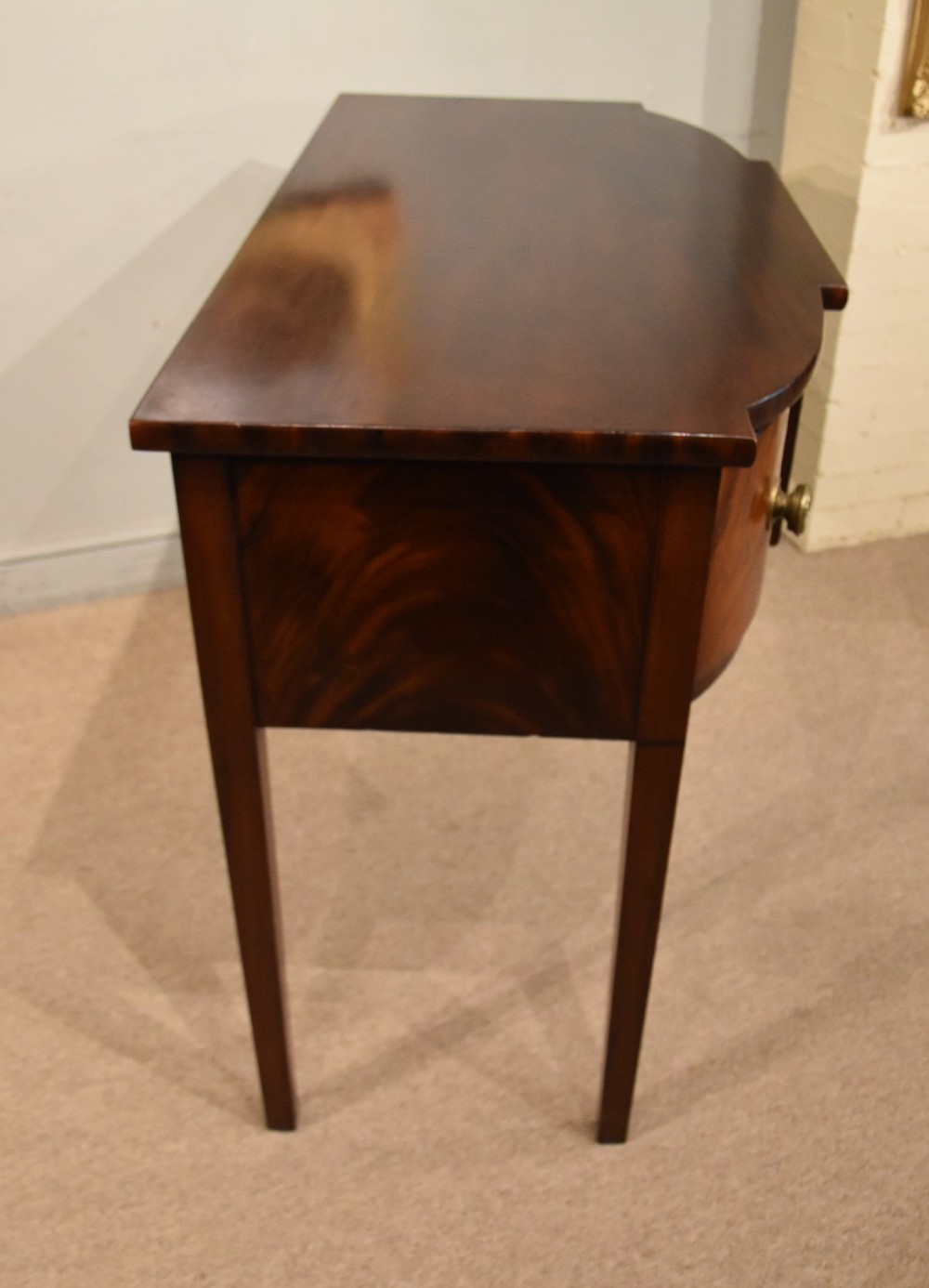 A Regency period flame mahogany bowfront sideboard featuring a central drawer flanked by two curved doors, set on tapered square legs with round brass handles. View VII