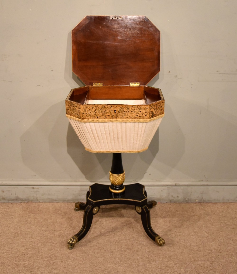 A Regency ebonised and gilt octagonal sewing table featuring detailed penwork on the lid, a pleated cream fabric basket beneath, and a central column resting on swept legs with brass castors. View II
