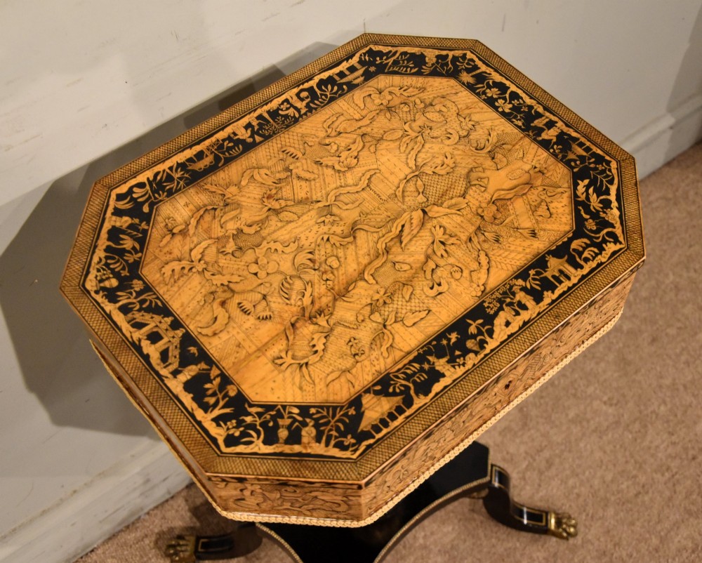 A Regency ebonised and gilt octagonal sewing table featuring detailed penwork on the lid, a pleated cream fabric basket beneath, and a central column resting on swept legs with brass castors. View V
