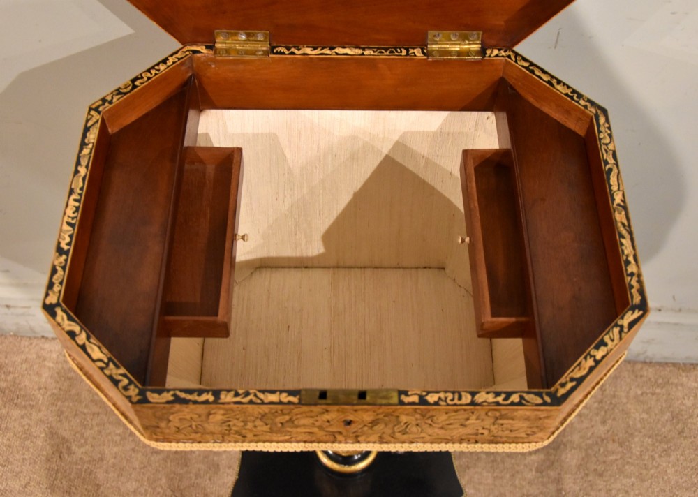 A Regency ebonised and gilt octagonal sewing table featuring detailed penwork on the lid, a pleated cream fabric basket beneath, and a central column resting on swept legs with brass castors. View VI