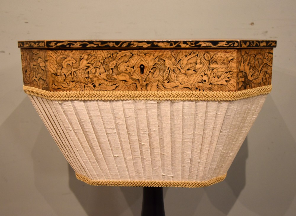 A Regency ebonised and gilt octagonal sewing table featuring detailed penwork on the lid, a pleated cream fabric basket beneath, and a central column resting on swept legs with brass castors. View VII