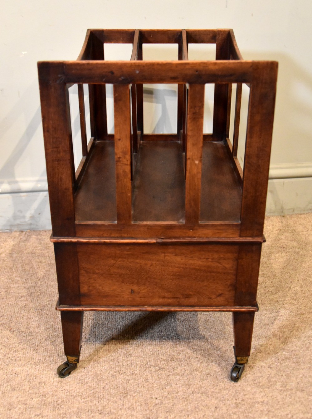 A 19th-century mahogany music canterbury featuring a curved four-compartment upper section above a single drawer with a central brass knob, raised on square tapered legs with brass castors. View V