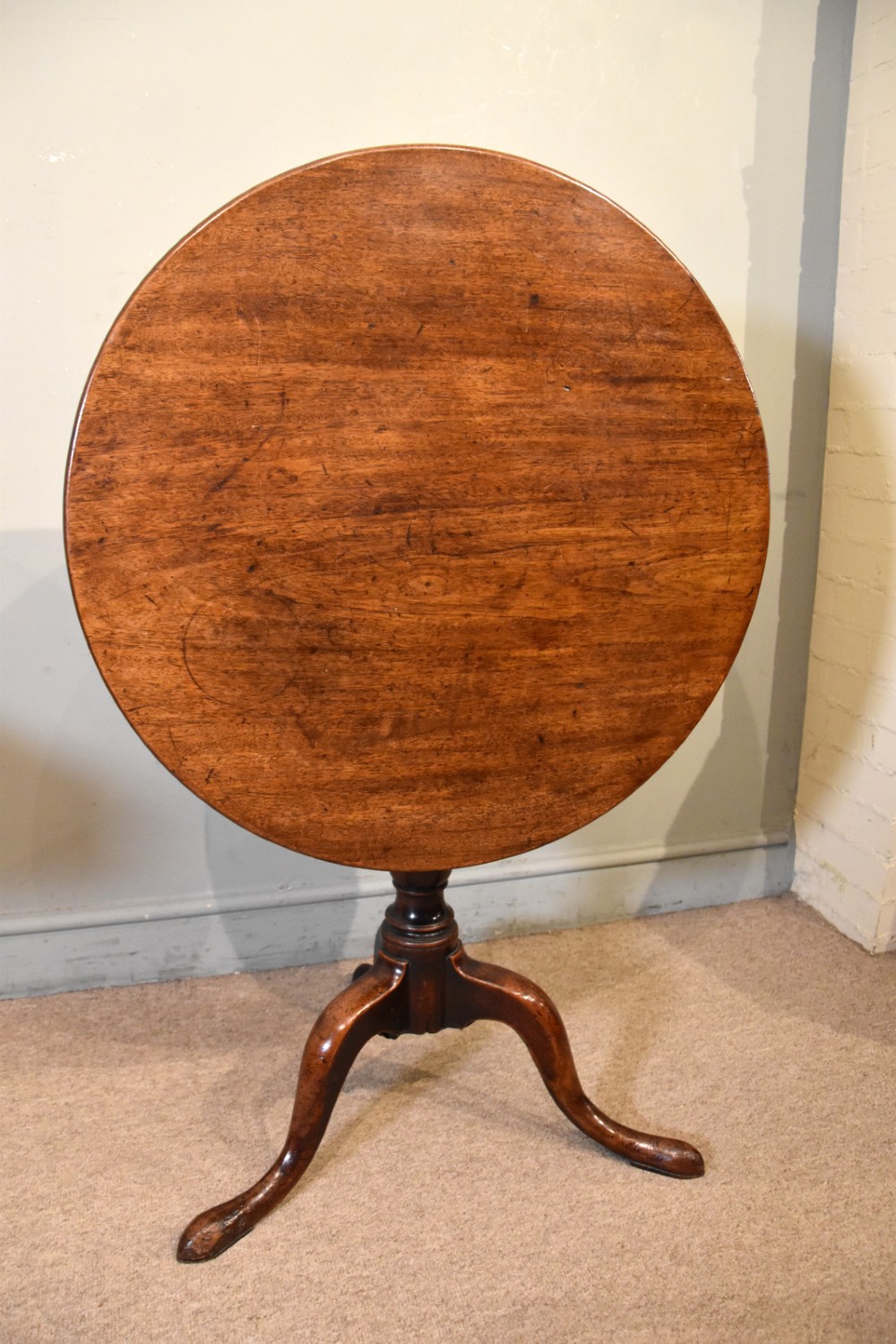 A George III mahogany tilt-top tripod table shown with its circular top tilted vertically on a turned stem and cabriole legs. View II