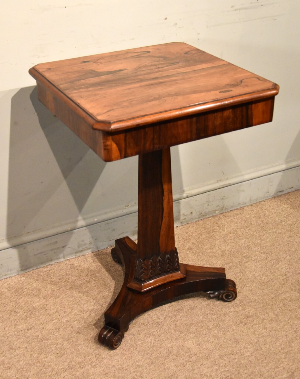 A William IV rosewood side table with a square top, plain frieze, tapered central pedestal, and a tri-form platform base on scroll feet. View IV
