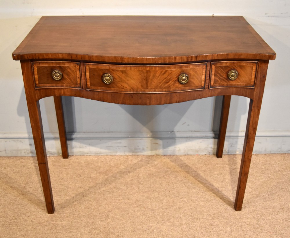 A George III mahogany dressing table featuring a serpentine front, three drawers with brass ring handles, crossbanded inlay, and four square tapered legs.