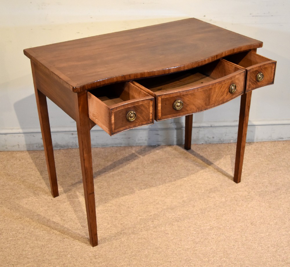 A George III mahogany dressing table featuring a serpentine front, three drawers with brass ring handles, crossbanded inlay, and four square tapered legs. View IV
