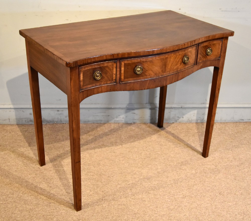 A George III mahogany dressing table featuring a serpentine front, three drawers with brass ring handles, crossbanded inlay, and four square tapered legs. View V