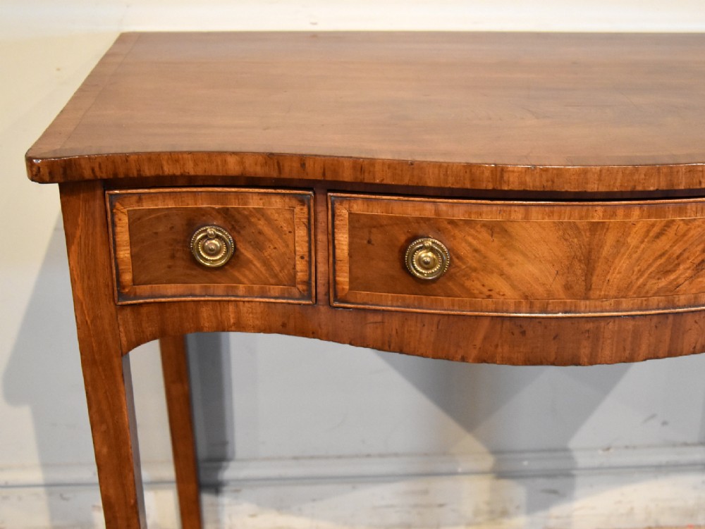 A George III mahogany dressing table featuring a serpentine front, three drawers with brass ring handles, crossbanded inlay, and four square tapered legs. View IX