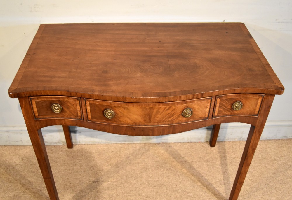A George III mahogany dressing table featuring a serpentine front, three drawers with brass ring handles, crossbanded inlay, and four square tapered legs. View X