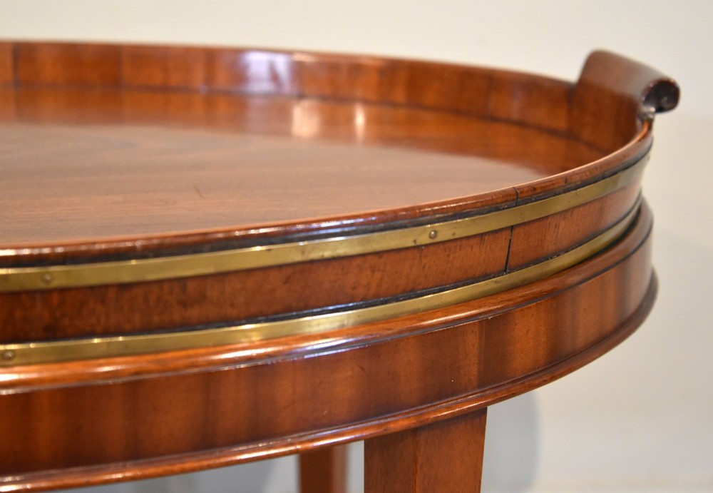 A late eighteenth-century oval mahogany tray table featuring a raised gallery edge, side cut-out handles, brass rim detailing, and tapered curved legs. View VI