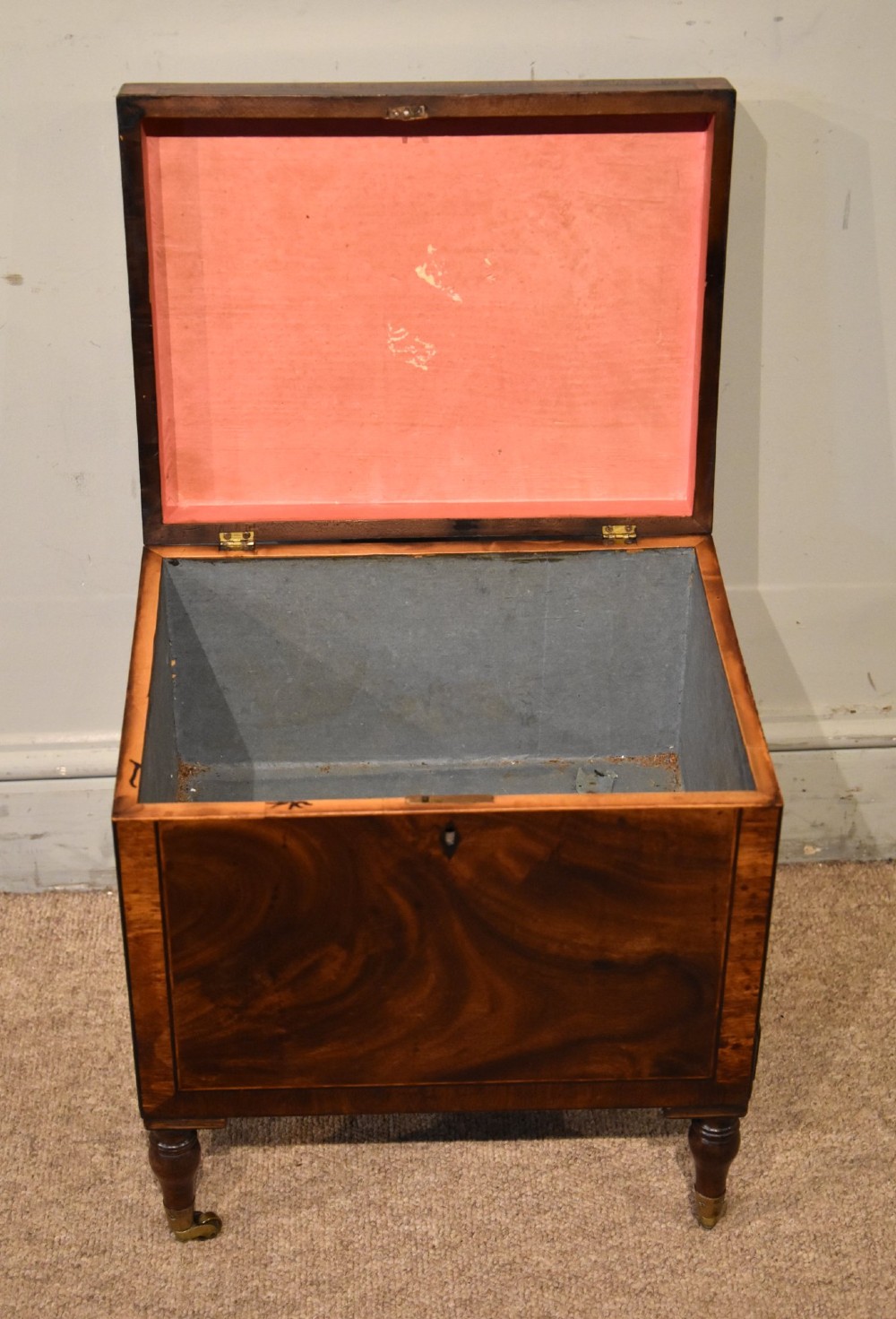 A Regency mahogany cellerette featuring flame timber veneers, crossbanded borders, a central keyhole, and turned legs on brass castors. View III