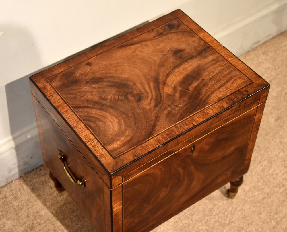 A Regency mahogany cellerette featuring flame timber veneers, crossbanded borders, a central keyhole, and turned legs on brass castors. View IV