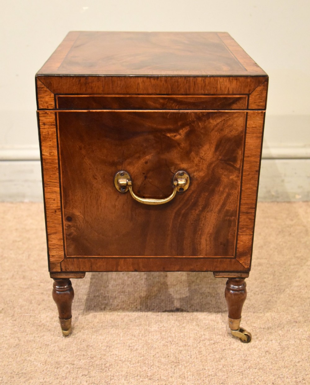 A Regency mahogany cellerette featuring flame timber veneers, crossbanded borders, a central keyhole, and turned legs on brass castors. View V