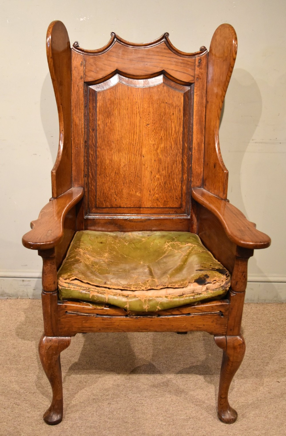 An 18th-century solid oak wingback armchair with a fielded panel back, shaped wings, cabriole front legs, and an aged green cushion.