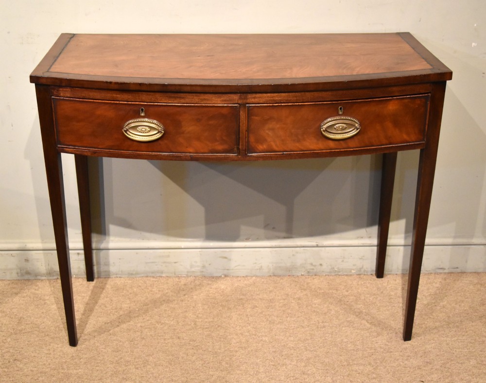 A George III bowfront mahogany serving table featuring two drawers with brass oval handles and slender tapered legs.