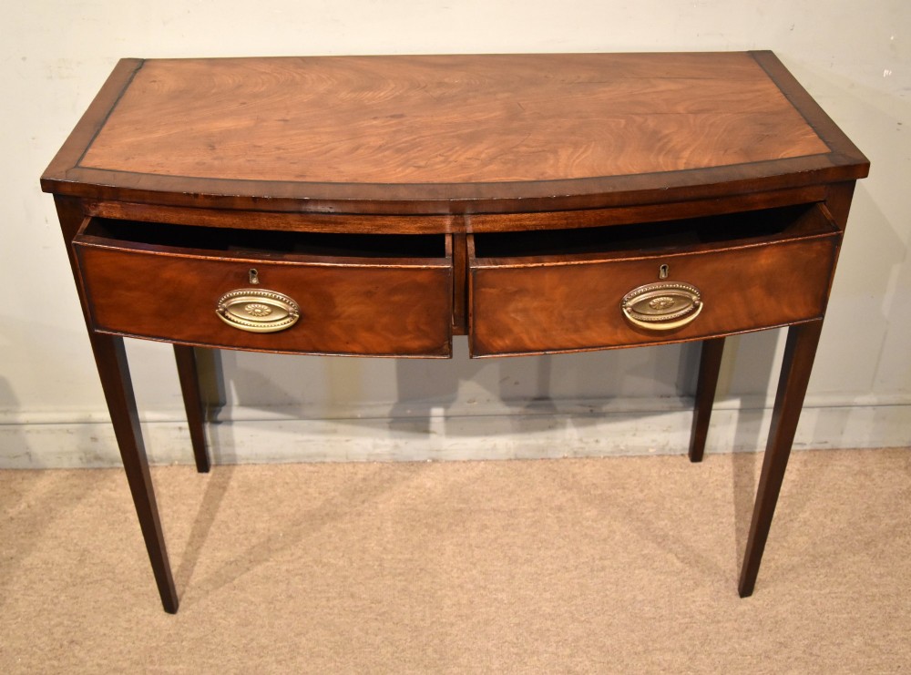 A George III bowfront mahogany serving table featuring two drawers with brass oval handles and slender tapered legs. View II