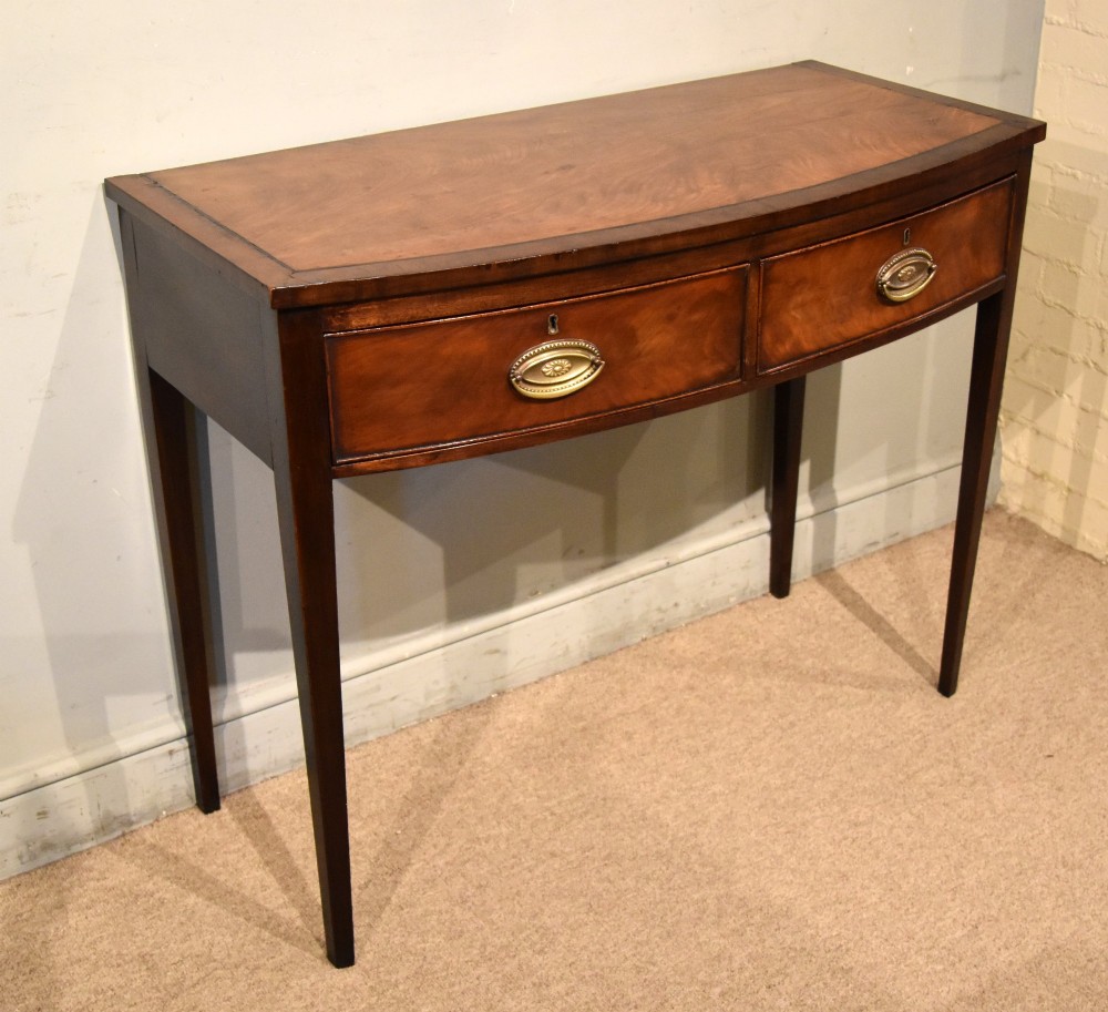 A George III bowfront mahogany serving table featuring two drawers with brass oval handles and slender tapered legs. View III