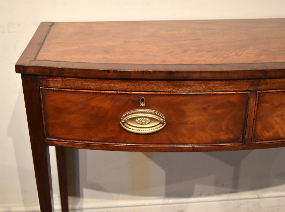 A George III bowfront mahogany serving table featuring two drawers with brass oval handles and slender tapered legs. View IV