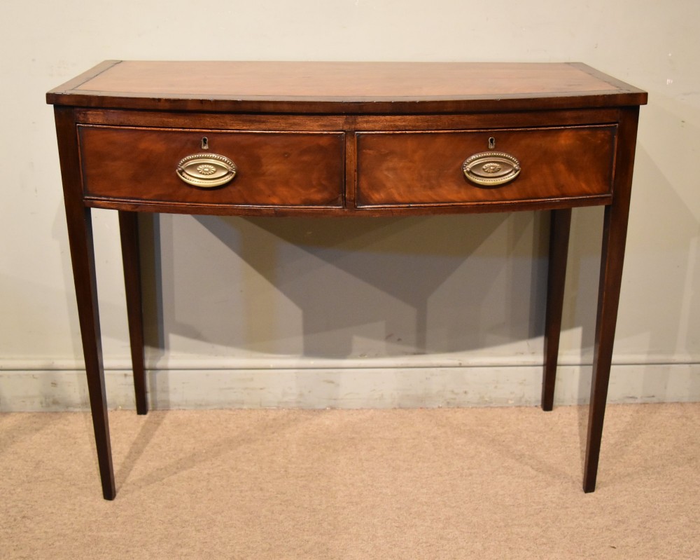 A George III bowfront mahogany serving table featuring two drawers with brass oval handles and slender tapered legs. View V