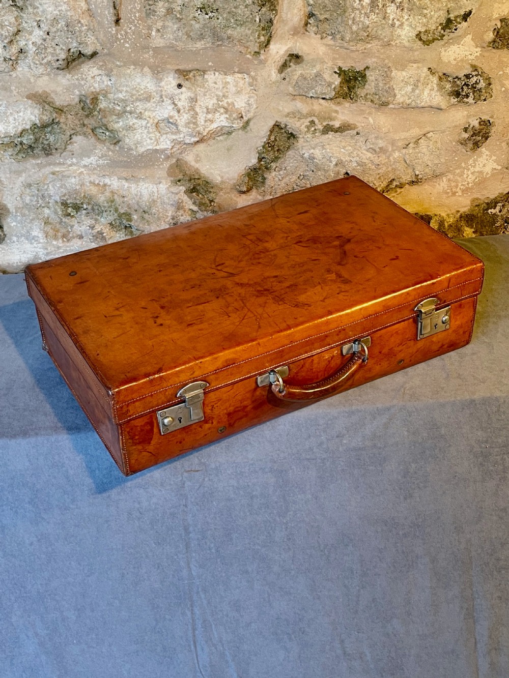 A rectangular chestnut brown leather suitcase featuring twin metal latches, hand-stitched edges, and a central leather carrying handle.