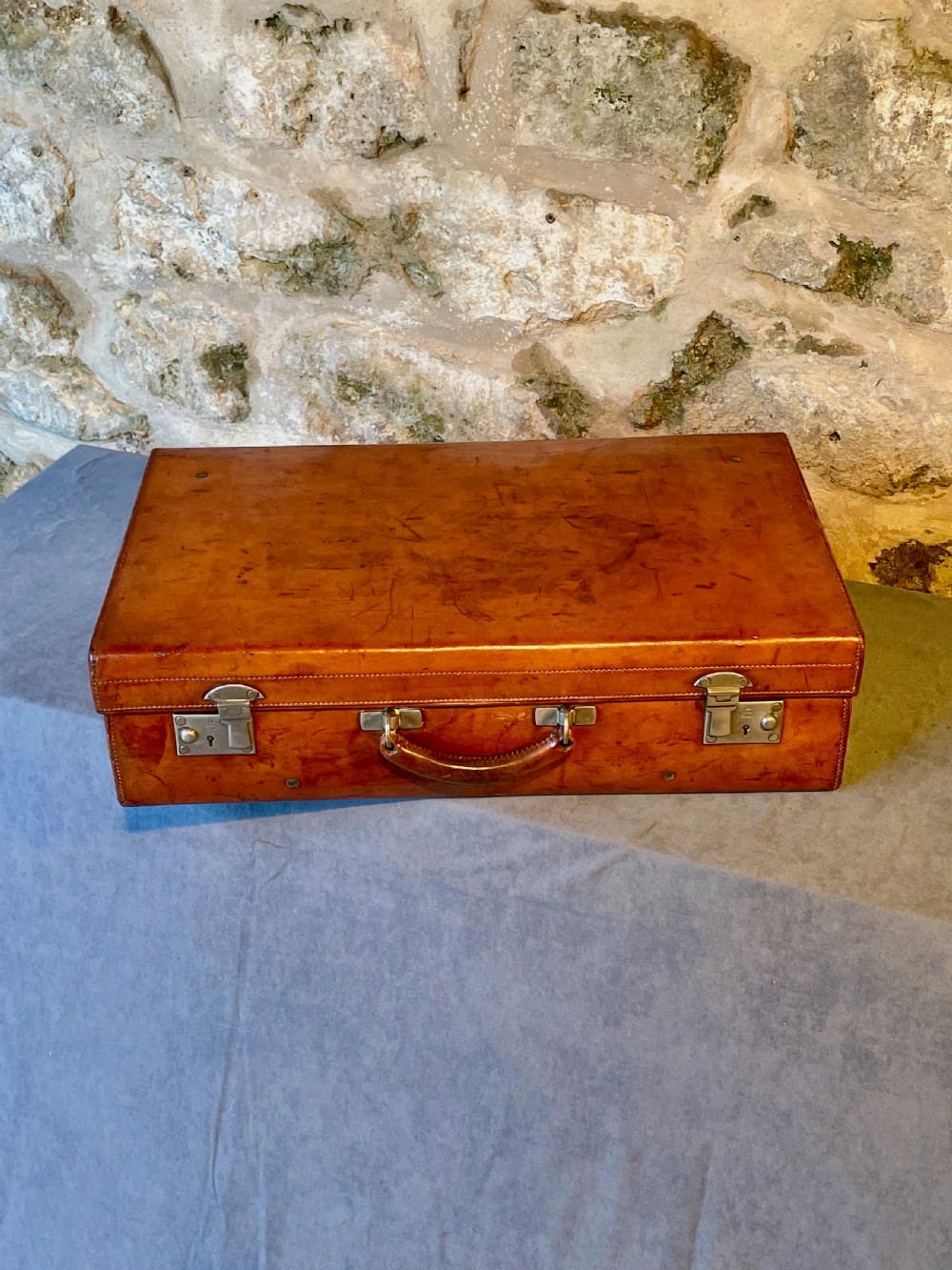 A rectangular chestnut brown leather suitcase featuring twin metal latches, hand-stitched edges, and a central leather carrying handle. View II