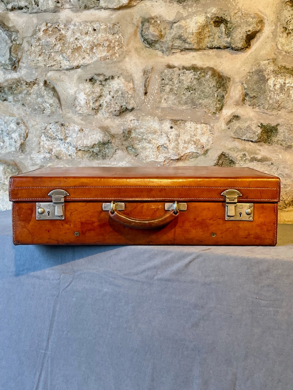 A rectangular chestnut brown leather suitcase featuring twin metal latches, hand-stitched edges, and a central leather carrying handle. View III