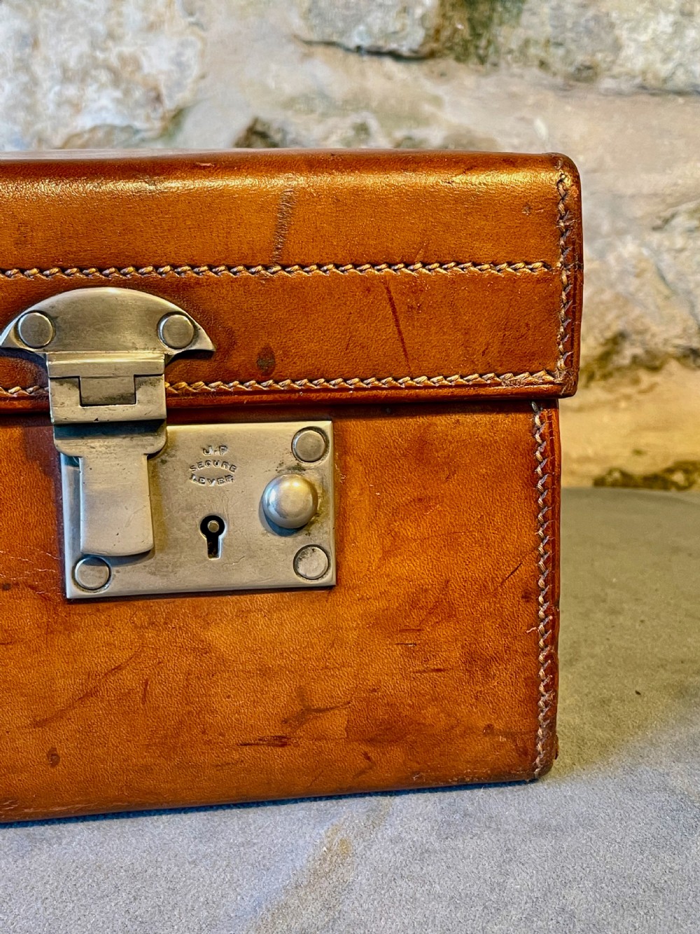 A rectangular chestnut brown leather suitcase featuring twin metal latches, hand-stitched edges, and a central leather carrying handle. View IV
