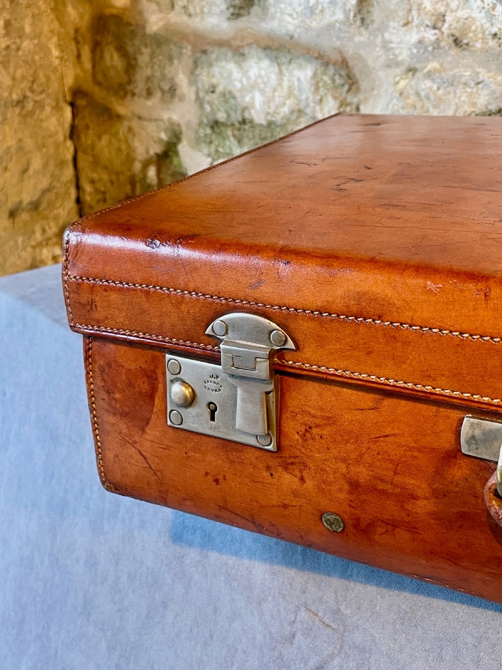 A rectangular chestnut brown leather suitcase featuring twin metal latches, hand-stitched edges, and a central leather carrying handle. View V