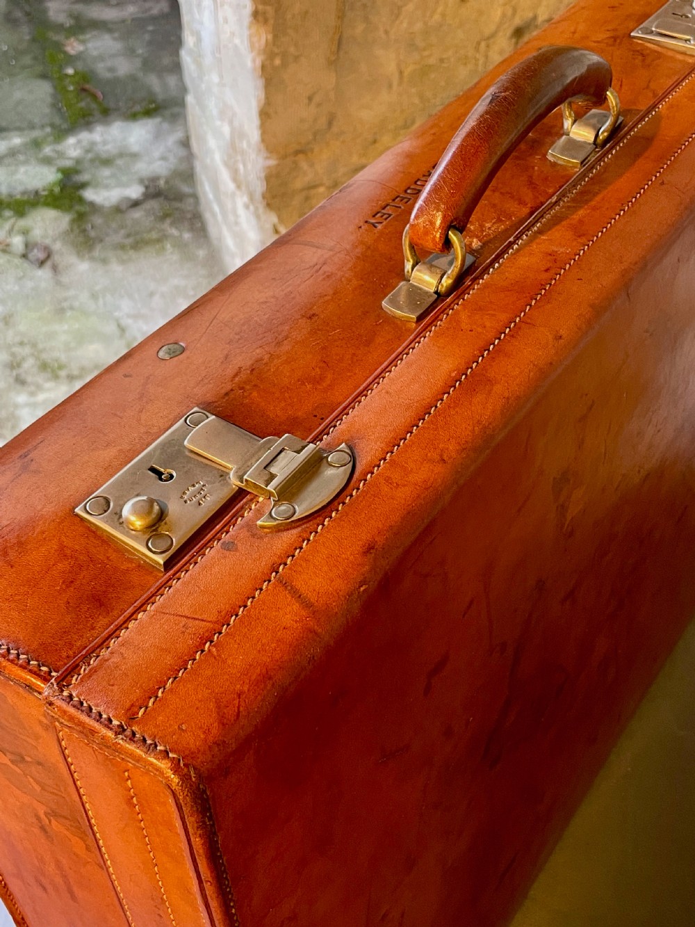 A rectangular chestnut brown leather suitcase featuring twin metal latches, hand-stitched edges, and a central leather carrying handle. View VI
