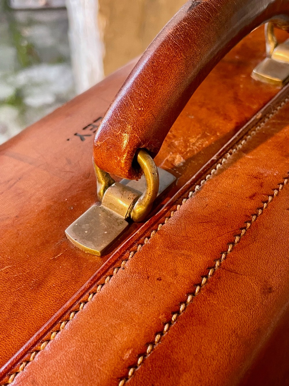 A rectangular chestnut brown leather suitcase featuring twin metal latches, hand-stitched edges, and a central leather carrying handle. View VII