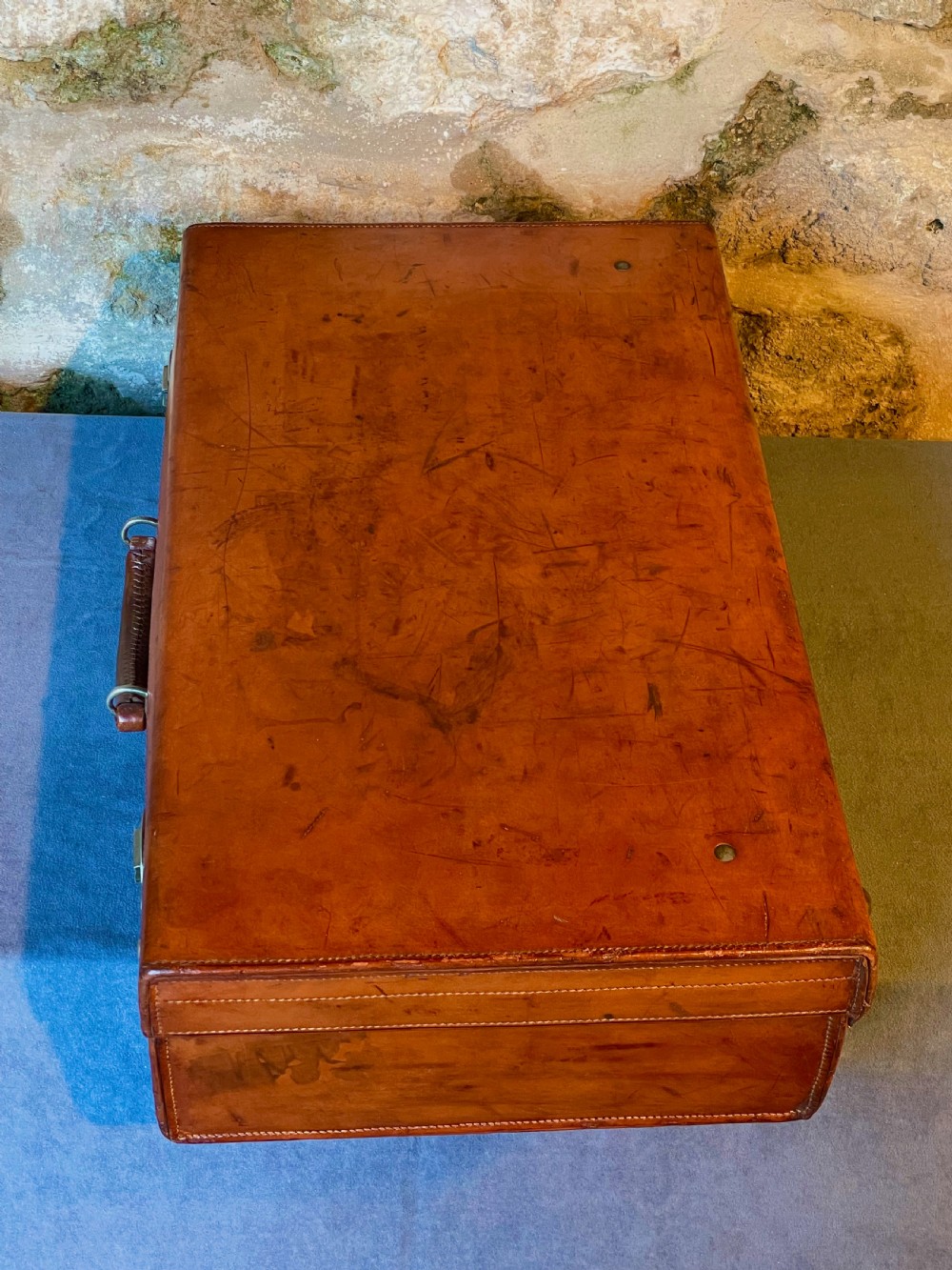 A rectangular chestnut brown leather suitcase featuring twin metal latches, hand-stitched edges, and a central leather carrying handle. View VIII