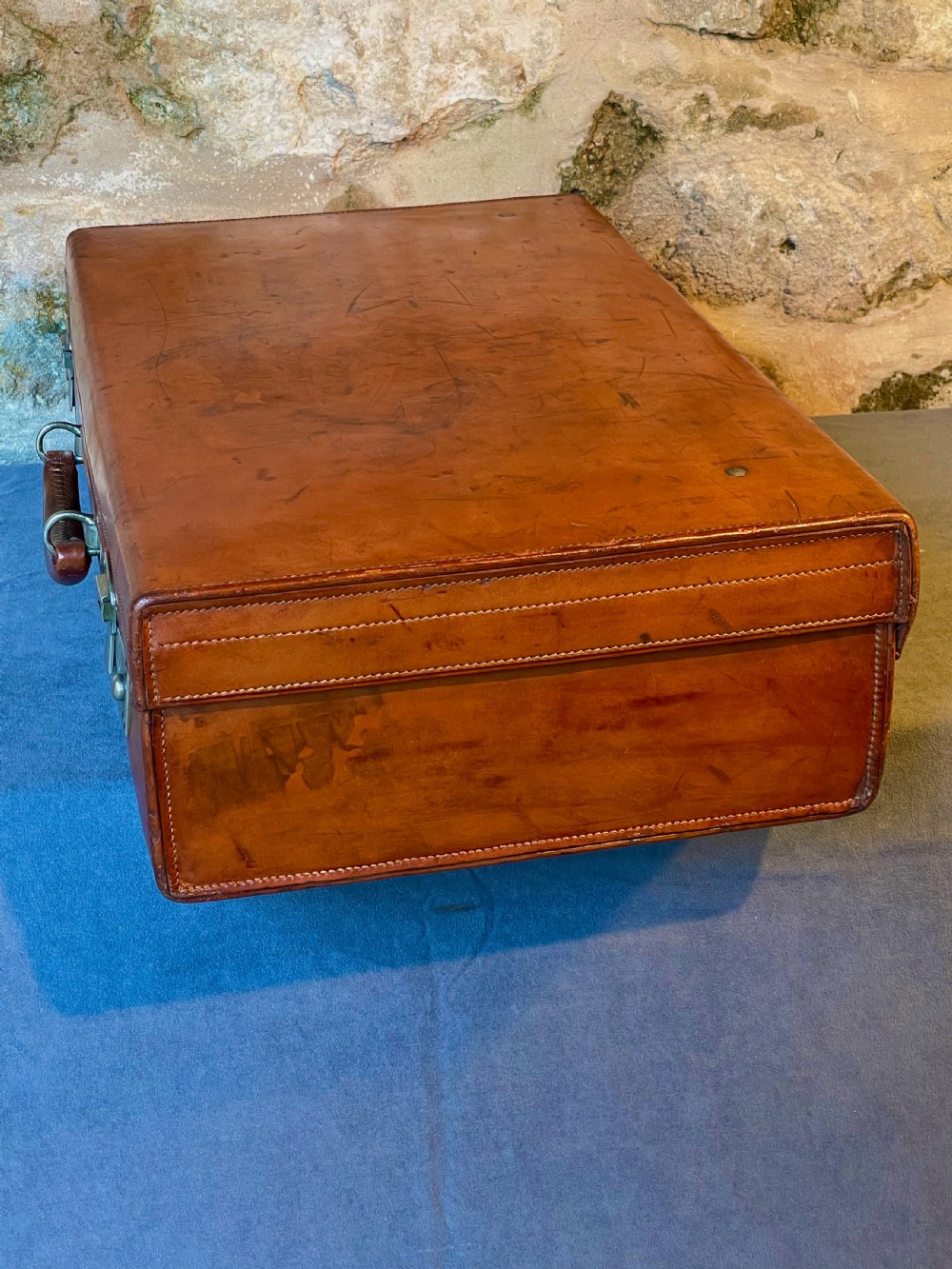 A rectangular chestnut brown leather suitcase featuring twin metal latches, hand-stitched edges, and a central leather carrying handle. View IX