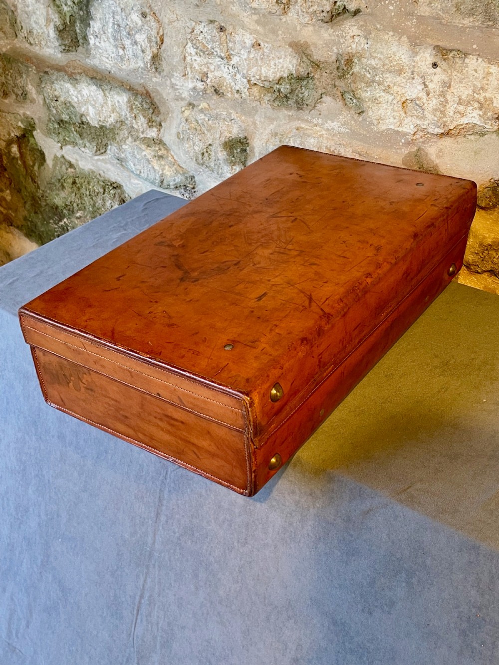 A rectangular chestnut brown leather suitcase featuring twin metal latches, hand-stitched edges, and a central leather carrying handle. View X