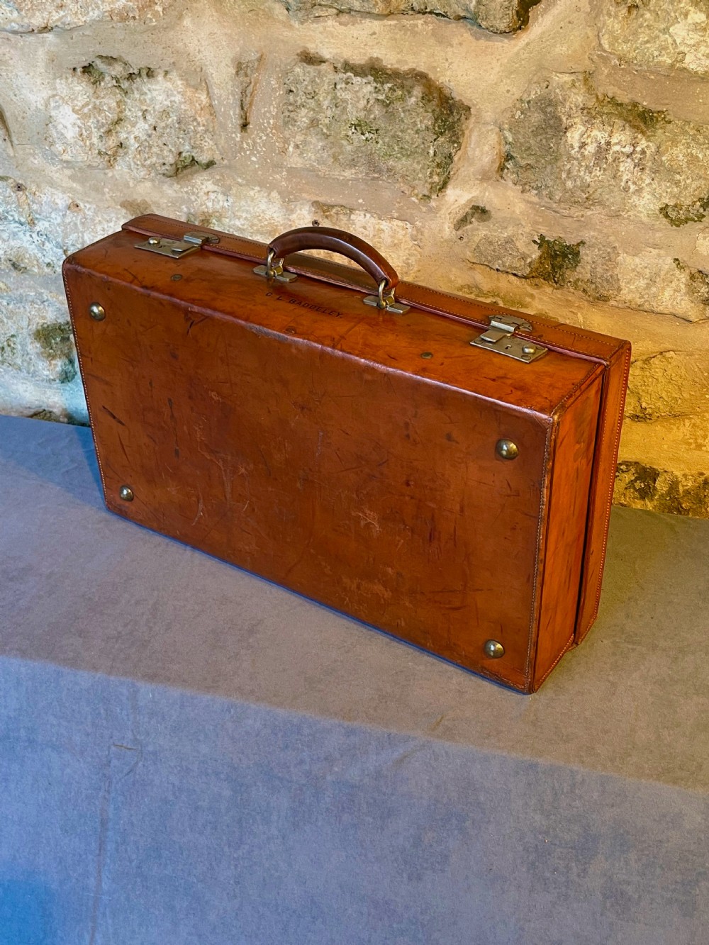 A rectangular chestnut brown leather suitcase featuring twin metal latches, hand-stitched edges, and a central leather carrying handle. View XI