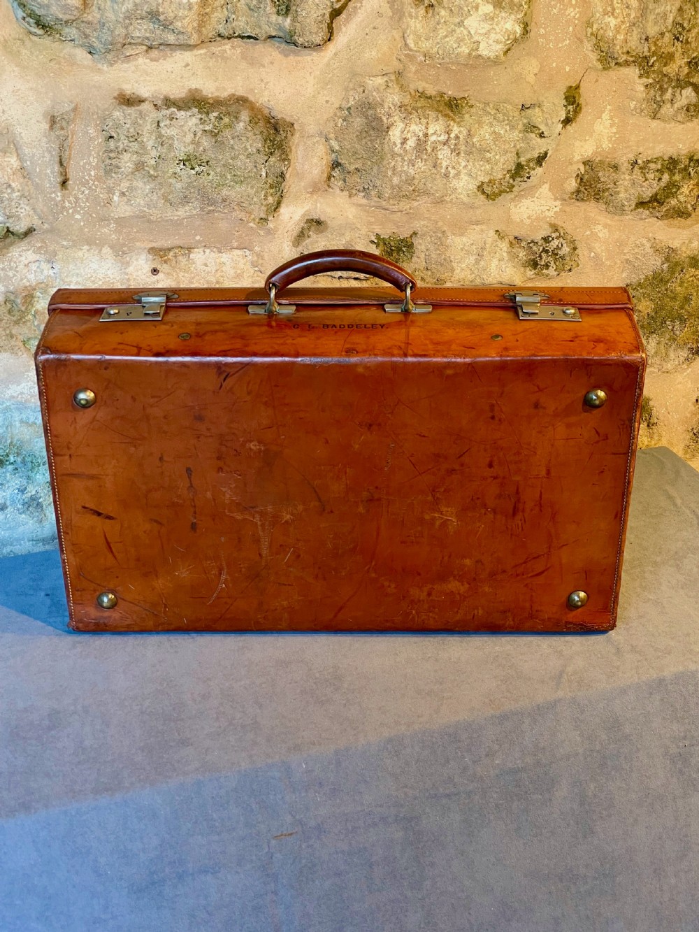 A rectangular chestnut brown leather suitcase featuring twin metal latches, hand-stitched edges, and a central leather carrying handle. View XII