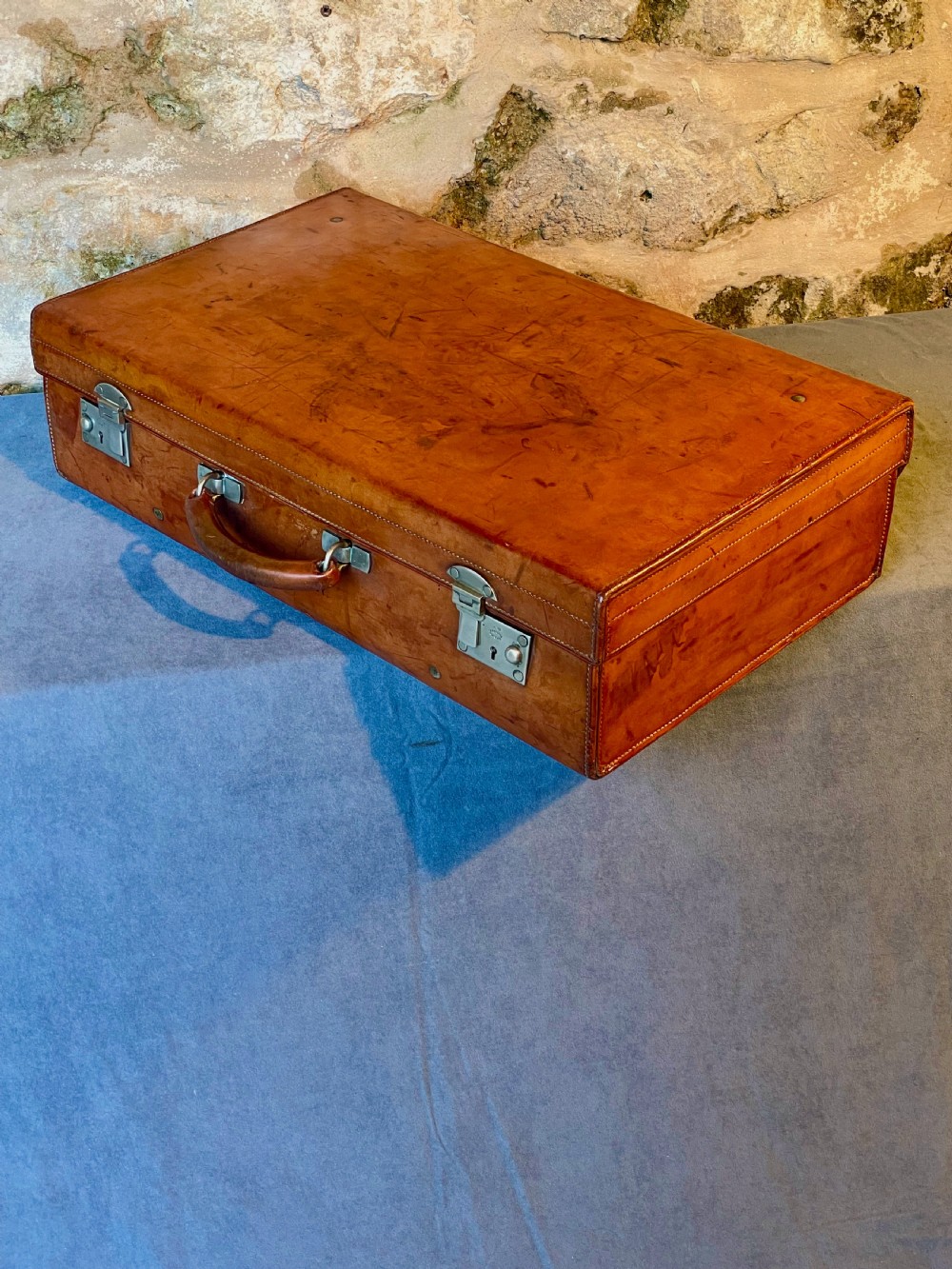 A rectangular chestnut brown leather suitcase featuring twin metal latches, hand-stitched edges, and a central leather carrying handle. View XIII