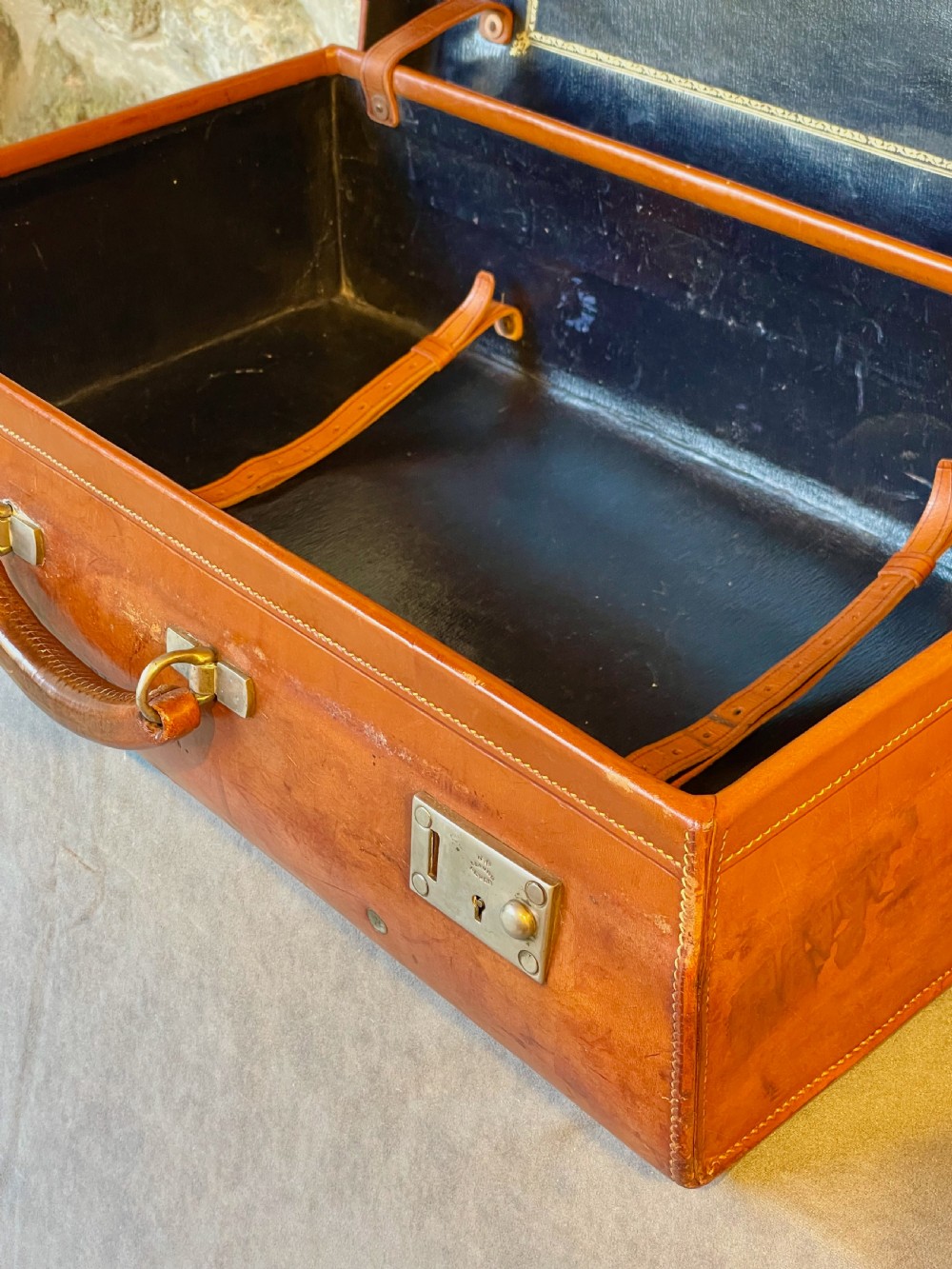 A rectangular chestnut brown leather suitcase featuring twin metal latches, hand-stitched edges, and a central leather carrying handle. View XVI