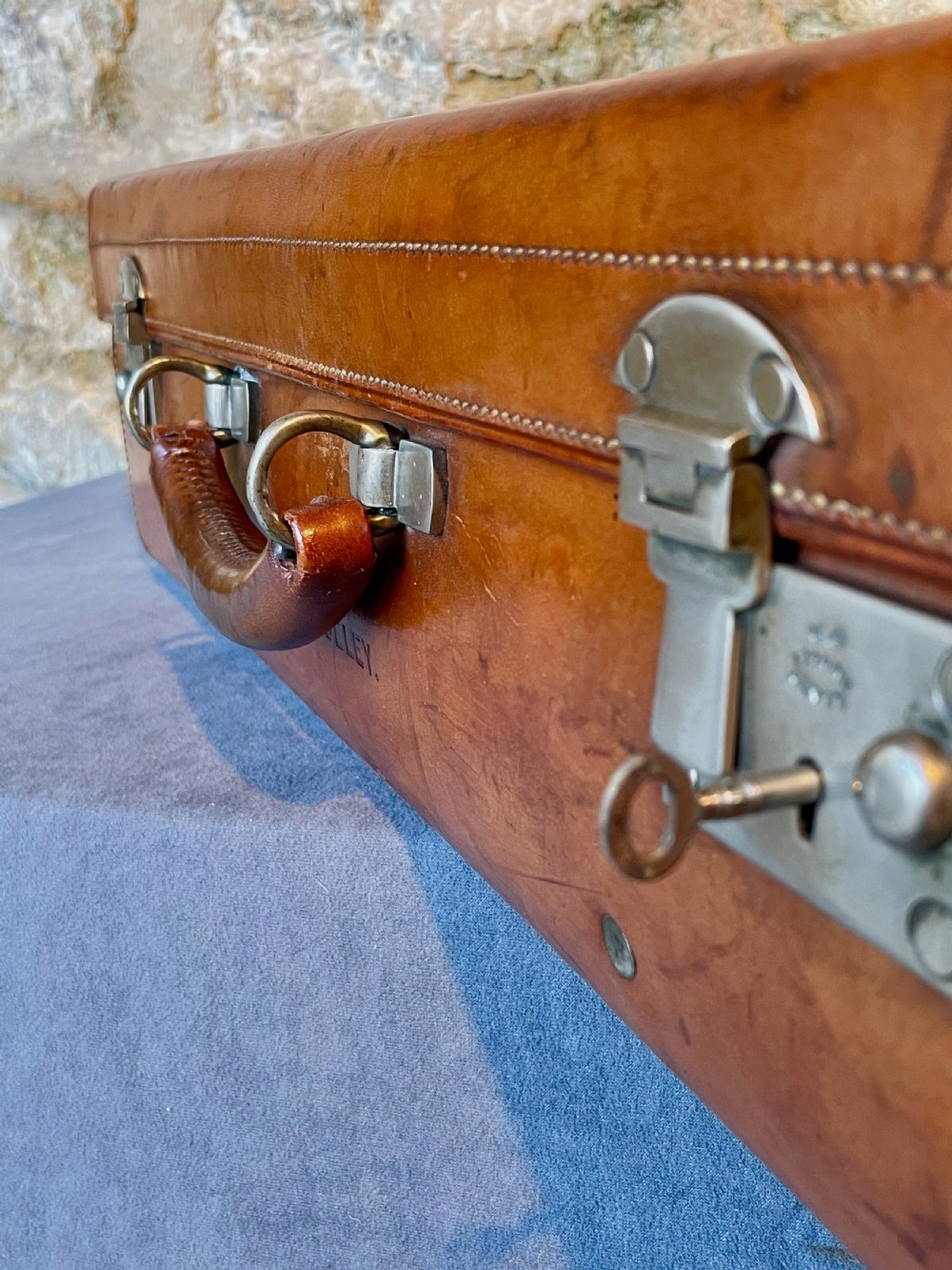A rectangular chestnut brown leather suitcase featuring twin metal latches, hand-stitched edges, and a central leather carrying handle. View XXII
