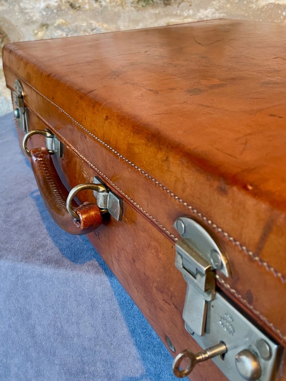 A rectangular chestnut brown leather suitcase featuring twin metal latches, hand-stitched edges, and a central leather carrying handle. View XXIII