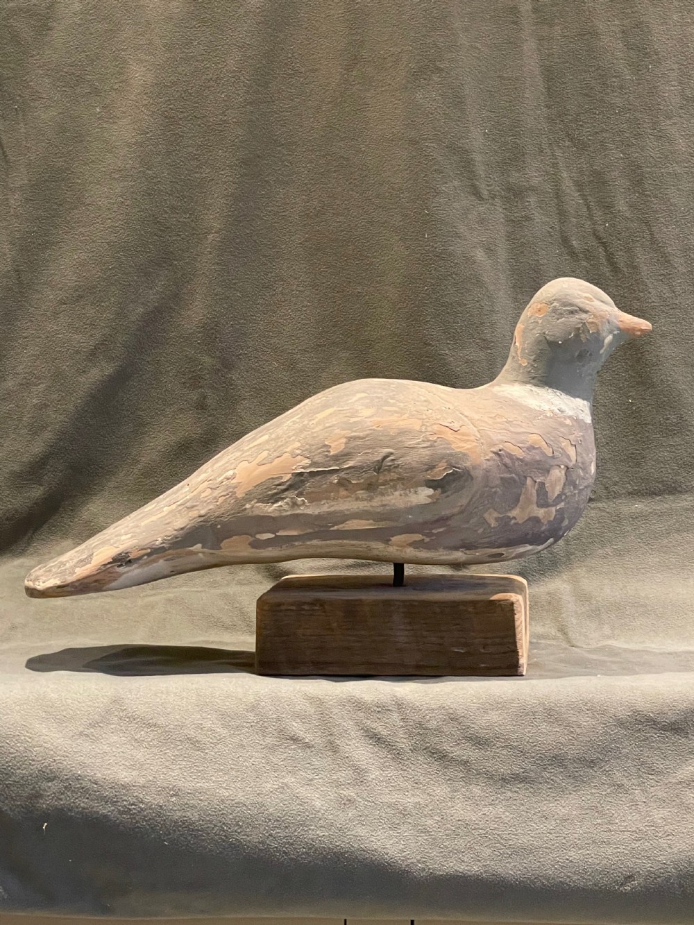 A hand-carved wooden pigeon decoy featuring weathered grey paint, mounted on a small rectangular wooden block.