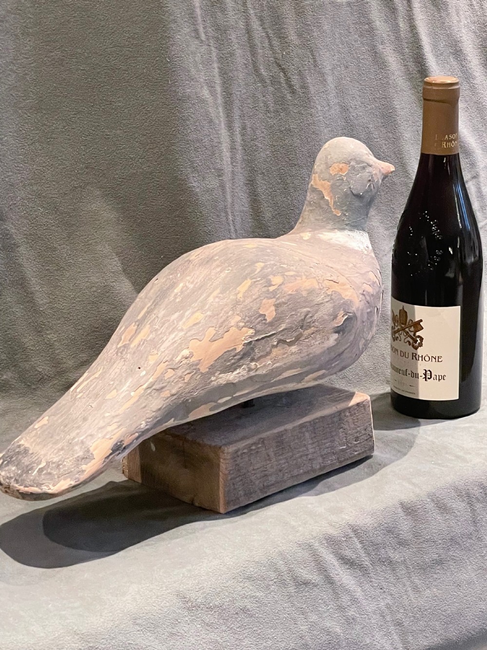 A hand-carved wooden pigeon decoy featuring weathered grey paint, mounted on a small rectangular wooden block. View IV