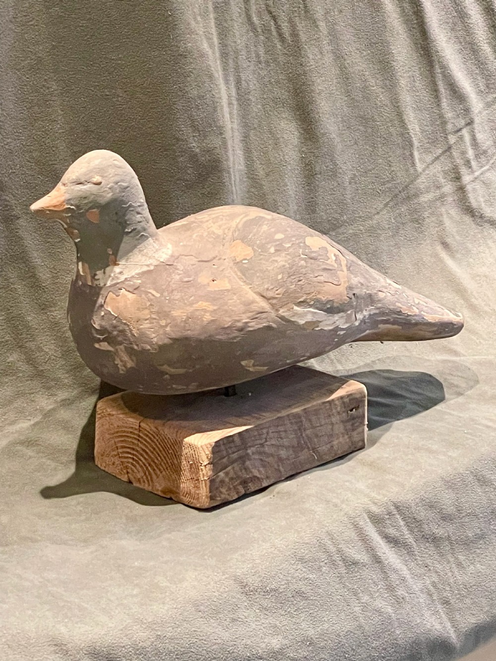 A hand-carved wooden pigeon decoy featuring weathered grey paint, mounted on a small rectangular wooden block. View VI